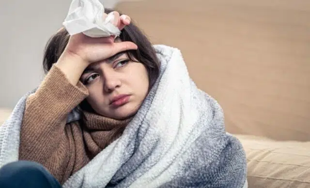 A sick young woman is sitting at home, wrapped in a blanket.