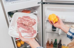 cropped view of woman holding frozen meat and bell pepper near open fridge with fresh food on