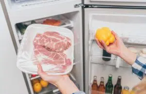 cropped view of woman holding frozen meat and bell pepper near open fridge with fresh food on