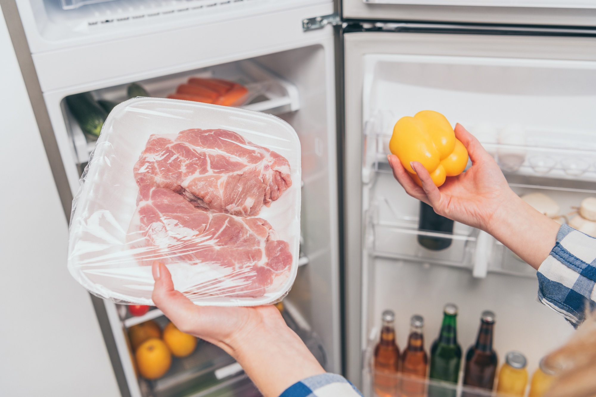 cropped view of woman holding frozen meat and bell pepper near open fridge with fresh food on