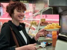 A woman in a black jacket smiles as she receives a plate of food from a man behind a counter.