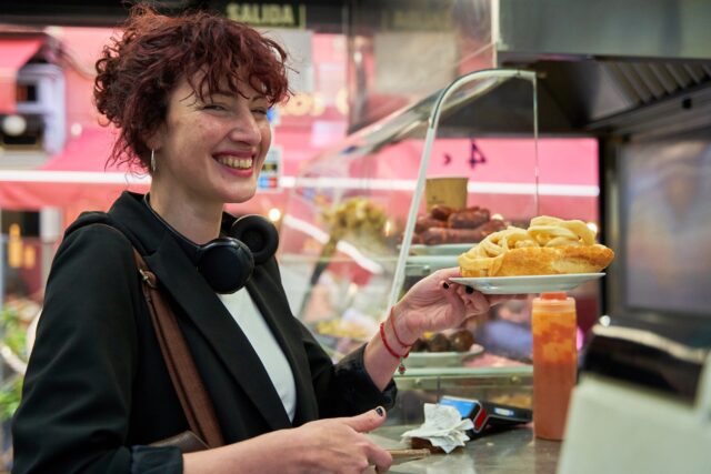 A woman in a black jacket smiles as she receives a plate of food from a man behind a counter.