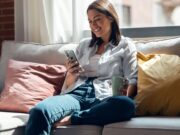 Pretty young smiling woman using mobile phone sitting on a couch at home.