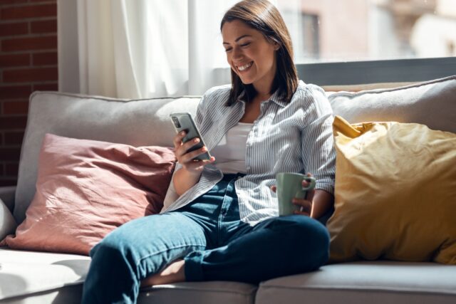 Pretty young smiling woman using mobile phone sitting on a couch at home. Pretty young smiling woman using mobile phone sitting on a couch at home.